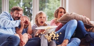 Frightened Family Sitting On Sofa At Home Watching Horror Movie On Tv With Popcorn