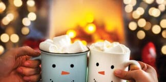 Mom And Child Are Relaxing Together On A Cozy Winter Evening By The Fireplace, Close Up Of Two Hands With Snowman Face Cup Of Hot Cocoa With Marshmallows. Christmas Holidays, Happy Moments At Home.